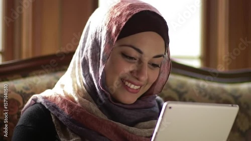 An Islamic woman, with a joyful smile, is engaged in online browsing and social media from her couch. The portrait captures a female wearing a hijab, using a digital device