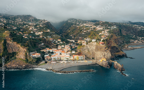 Aerial view of the coastal village nestled beneath the cliffs, where colorful buildings meet the dark, shimmering ocean, Ponta do Sol, Madeira, Portugal.