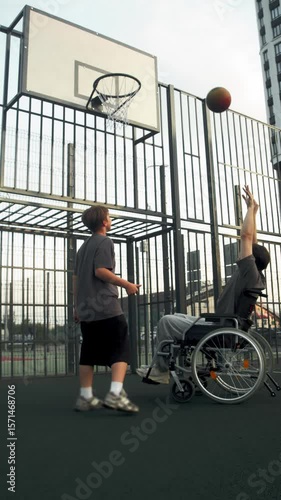 A young father, sitting in a wheelchair, plays basketball with his son. They manage to score points and are very happy together. Vertical.