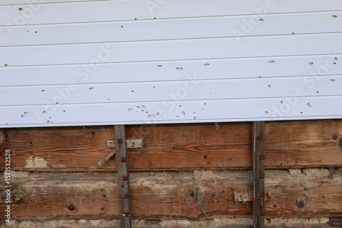 Quadro su tela Partially detached plastic siding reveals damaged wooden cladding and an old wall covered with numerous hail marks