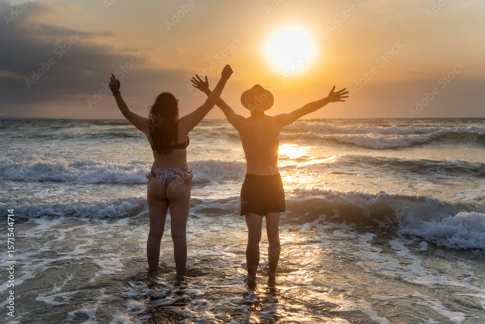 Fototapeta premium Couple enjoying golden sunrise on the beach, raising arms in gratitude