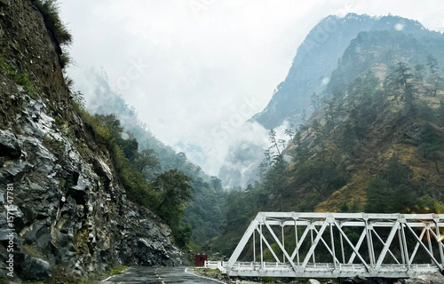 Mountain Road with White Iron Bridge surrounded by Steep Rocky Cliffs and Pine Forests on a misty day , Image of Dong Arunachal Pradesh  
