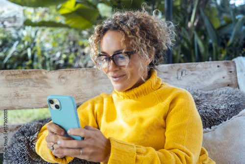Attractive woman in yellow sweater uses cell phone and relaxes alone in the garden sitting on a sofa. Portrait of serene female writing on cell phone. Mature young people relaxing
