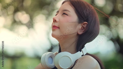 Close-up of peaceful asian woman with headphones looking up calmly while enjoying gentle breeze in city park – wellness lifestyle, good health and positive mental wellbeing in relaxing urban nature
