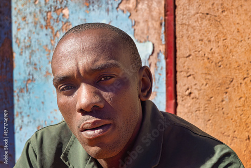africa village, single african man headshot, in the township , tough manly look, standing outdoors in the yard in front of the house door