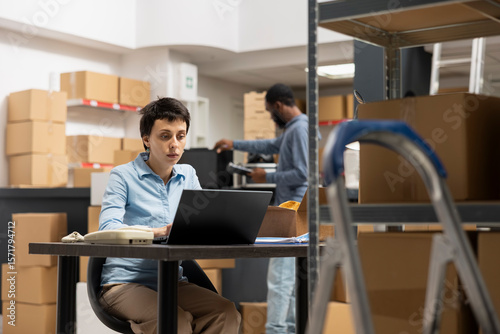 Female business owner works on a laptop in a local storehouse, focusing on shipping logistics for custom orders and local express delivery service. Storage room retail activity.