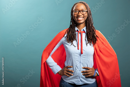 African american woman in red cape and blue shirt, hands on waist, smiling brightly at camera. Black female model stands in a heroic pose, portraying a confident corporate superhero.