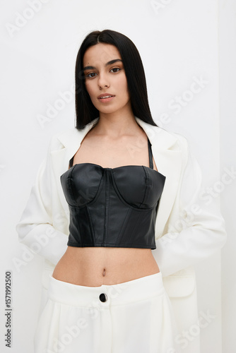 Young woman wearing a black leather corset top and white blazer, posing confidently with straight long hair against a plain white background.