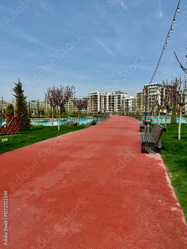 Red running path in a modern city park on a sunny day. Urban recreational area with apartment buildings and a blue sky.