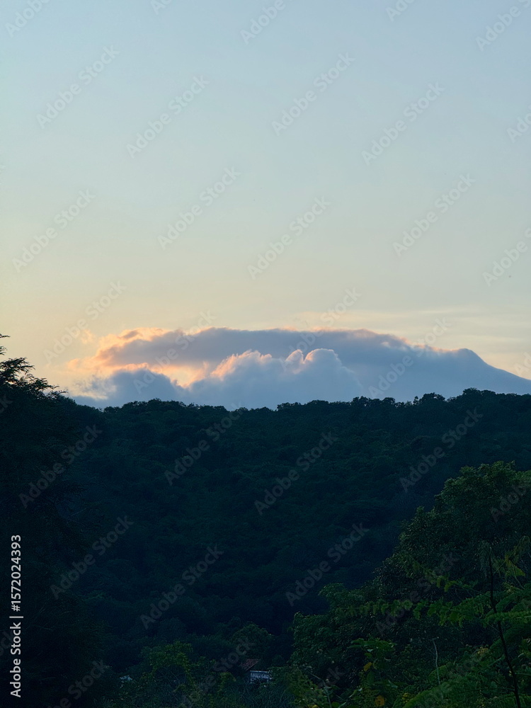 Naklejka premium Peaceful mountain landscape at dusk. Glowing clouds from the sunset surround a distant peak above a lush green forest.