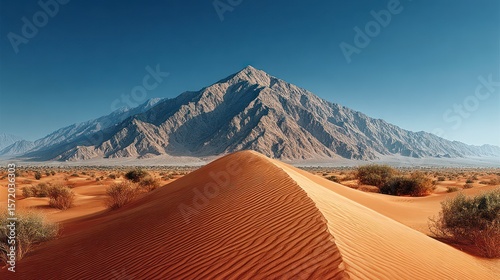 Rippled desert sand dunes stretch towards a large mountain range under a clear blue sky
