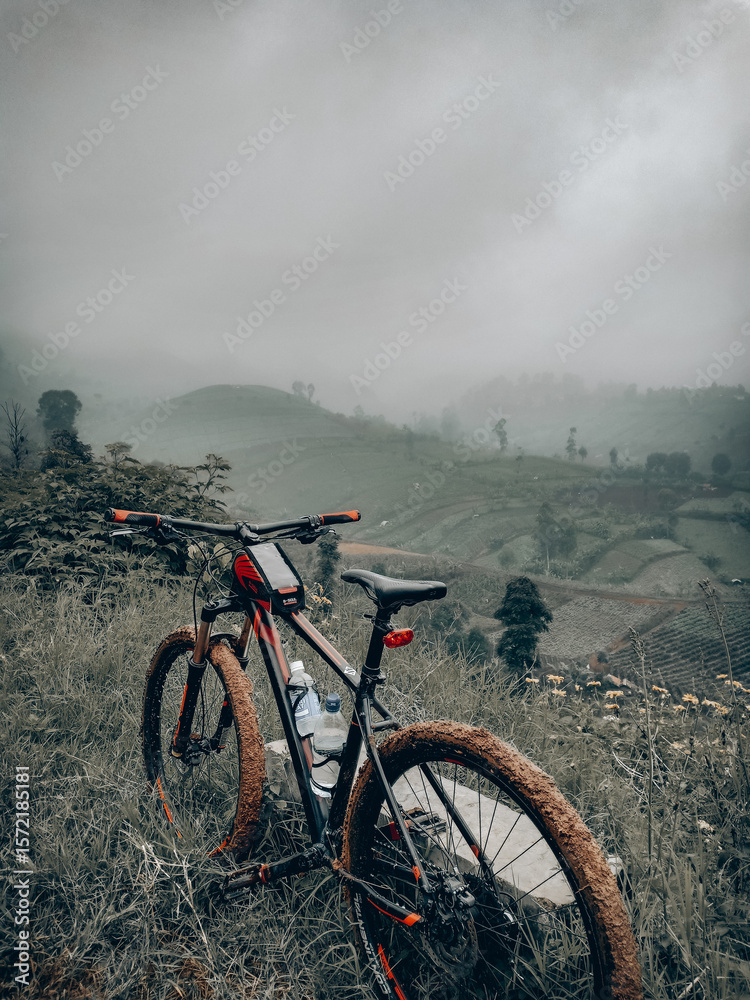 Obraz premium A scenic view from a bridge shows a mountain bike resting against a red and black railing. In the background beautiful rice field views under a foggy weather.