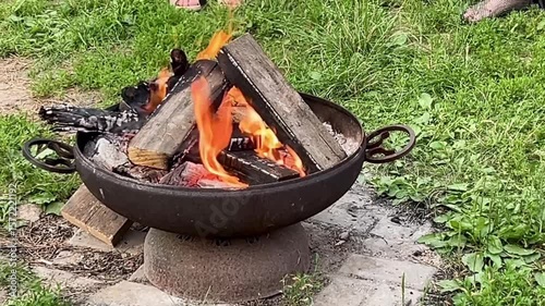 Rusty fire pit with crackling firewood creating a warm, inviting atmosphere in a backyard setting. Vibrant flames against a lush green backdrop, perfect for outdoor gatherings.
