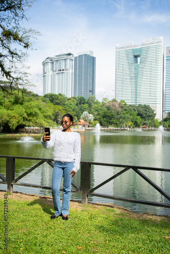 Photography Young woman taking selfie in a park with skyscrapers and lake in kuala lumpur