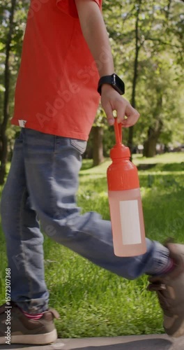 Junior schoolboy in casual clothes walks slowly through city park and carries bright water bottle in hand. Child holds vibrant plastic bottle. Vertical shot