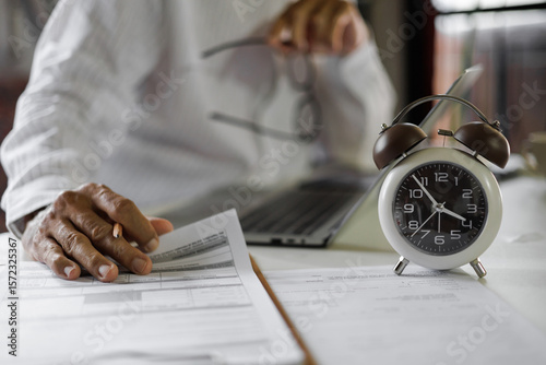selective focus on alarm clock, asian businessman review business document using laptop working on table before work time end in a day,  time management concept, routine work, office hour