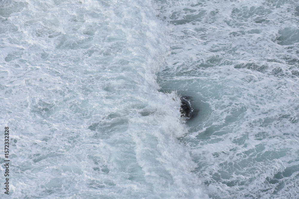 Fototapeta premium Powerful waves crash over a dark rock beneath the swirling turquoise waters of Nazaré, Portugal — nature’s raw energy frozen in motion.