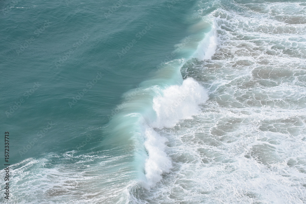 Fototapeta premium A powerful turquoise wave crashes onto the shore, captured in stunning detail from above. The seafoam swirls in vibrant white against the crystal-clear blue water