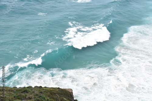 A powerful turquoise wave crashes onto the shore, captured in stunning detail from above. The seafoam swirls in vibrant white against the crystal-clear blue water