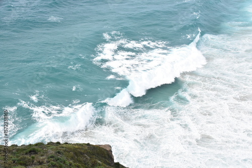 A powerful turquoise wave crashes onto the shore, captured in stunning detail from above. The seafoam swirls in vibrant white against the crystal-clear blue water