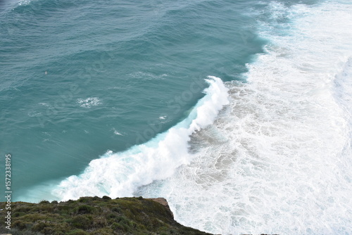A powerful turquoise wave crashes onto the shore, captured in stunning detail from above. The seafoam swirls in vibrant white against the crystal-clear blue water
