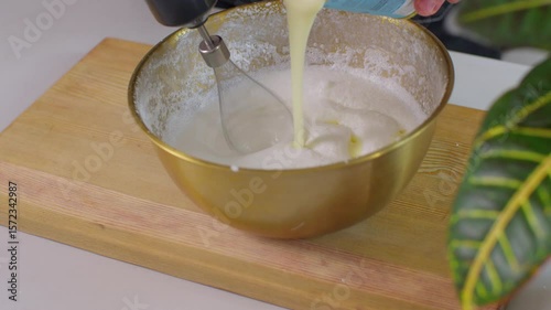 Adding condensed milk to whipped cream in a golden bowl and mixing with a hand mixer. Dessert preparation in home kitchen.