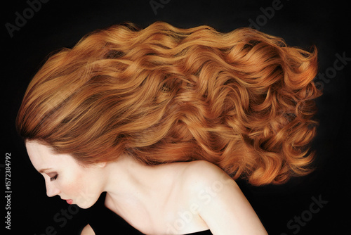 Studio portrait of beautiful young woman with long, curly red hair against black background
