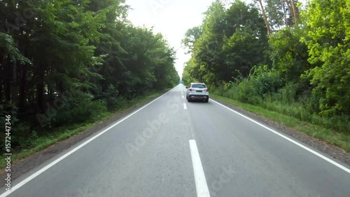 Aerial shot of car riding through forest rural road. White SUV driving at empty country route near wood on summer day. Follow to auto moving during journey or trip. Scenic landscape way. Slow motion