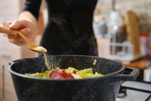 A person is adding spices to a pot filled with chopped vegetables and meat