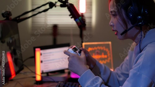 Young Asian female streamer playing video game live stream from home gaming room setup using controller headset Shows focused expression in vibrant lighting