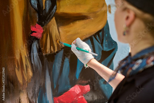 Focused view of adults hand painting mural outdoors. Gloved hand holds brush applying vivid red and blue hues on rough surface with precision and artistry