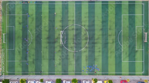 Top-down aerial view of children training and playing football on a striped green artificial turf soccer field.