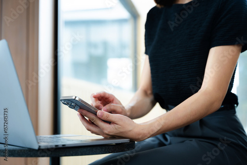 Asian businesswoman using smartphone and laptop in a modern workspace by a window, showcasing digital communication