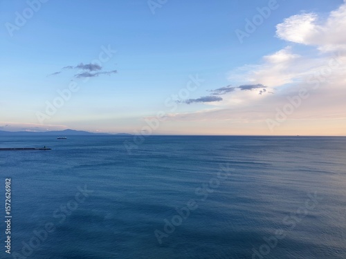 A calm ocean stretches to the horizon. A breakwater is to one side, with a boat framed by distant hills. The water is dark blue, and the clouds have the colours of sunset.