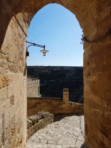 A stone archway opens onto a view of distant hills. A cobbled footpath with stairs lead down the hill and run beside a low stone wall. An old street light is seen against a blue sky.