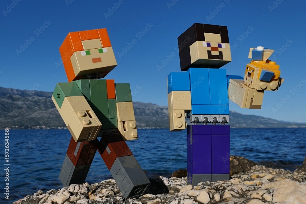 Obraz premium LEGO Minecraft Steve holding LEGO small and large Pufferfishes aquatic mobs, Alex standing next to him. Location rocky Croatian beach, bay water and Velebit mountain in background. Blue summer skies.
