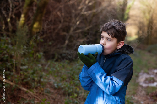 A young boy drinks from a blue water bottle while standing in a green forest, he is dressed warmly in outdoor clothing. Boy resting after running in forest and drinking water