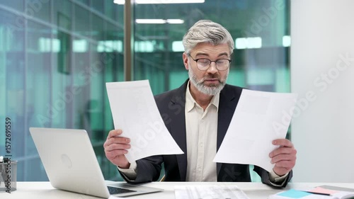 Confused disappointed mature businessman having difficulty with paperwork sitting at desk at workplace in business office. Puzzled worker in a suit looks at documents and cannot understand the problem