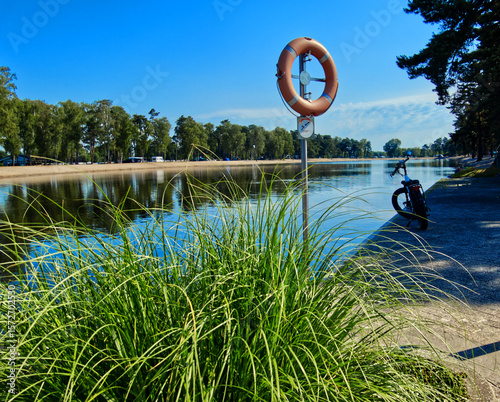 Electric bike resting beside lifebuoy on sandy lakeshore, pine trees framing sunny summer landscape