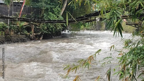 The river flows fast after the rain in the landscape. Rapid ripples crashed against the rocks on the surface, with green trees along the edges.