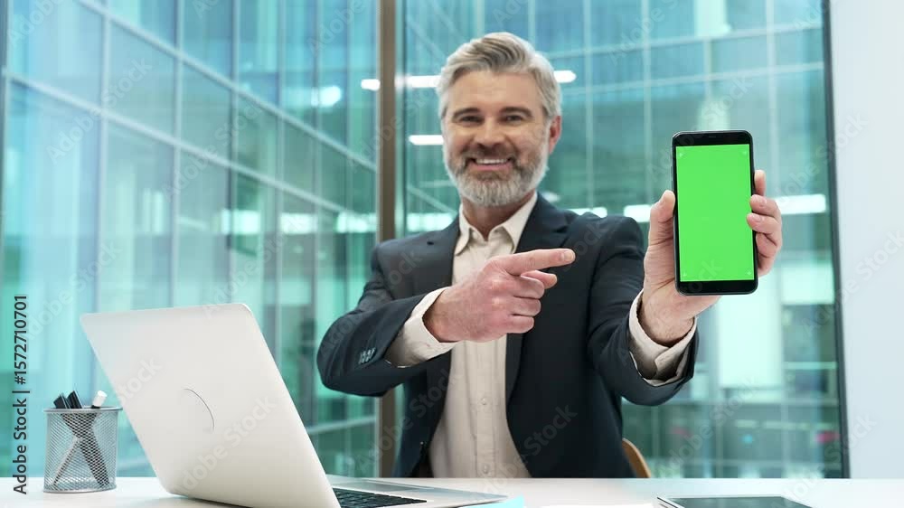 Happy businessman pointing finger at mobile phone with green screen sitting in business office. Color key, vertical template layout for advertising. Smiling male looking at camera showing smartphone