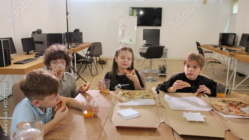 A group of children are seated around a table in a minimalist office space, enjoying slices of pizza and sipping on juice. Laughter fills the air as they engage in conversation.