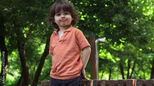 A young child with medium-long hair, wearing an orange polo shirt, stands in a green park or forest