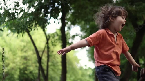 A young child with medium-long hair, wearing an orange polo shirt, stands in a green park or forest