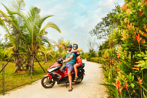 Funny authentic young happy caucasian couple pose on scooter together with palm trees, having fun driving red scooter on tropical road trip in southeast asia. Couple in love having fun travel in asia