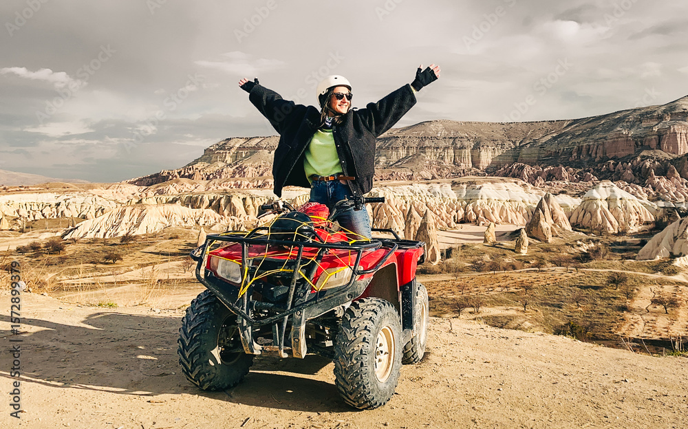 Obraz premium Goreme, Turkey - 26th december, 2024: happy joyful excited woman adventurer hands up sit on red squad over Cappadocia red valley. ATV squad group tour viewpoint. Travel Turkey in winter outdoors