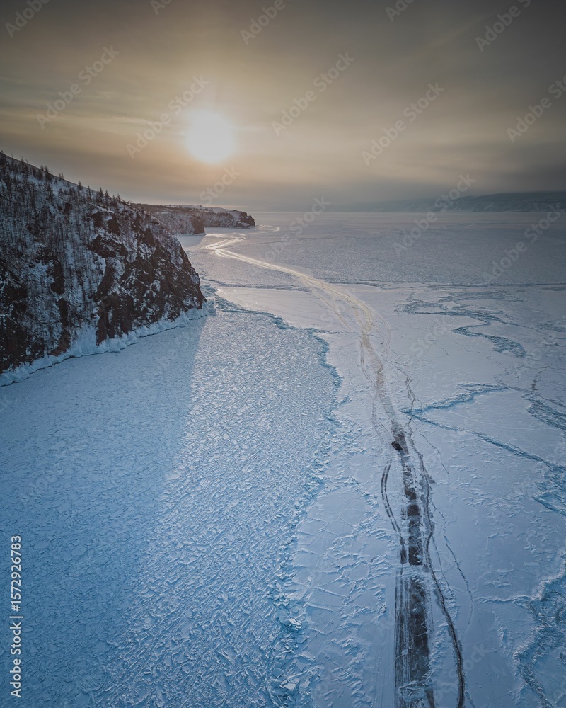 Obraz premium Frozen lake with cracked ice and sunset over cliffs.