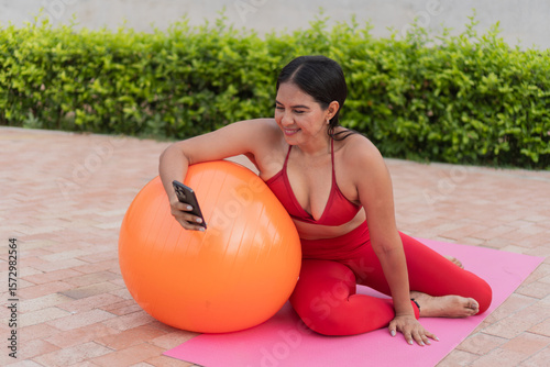 Latin woman using smartphone while doing pilates with an exercise ball outdoors