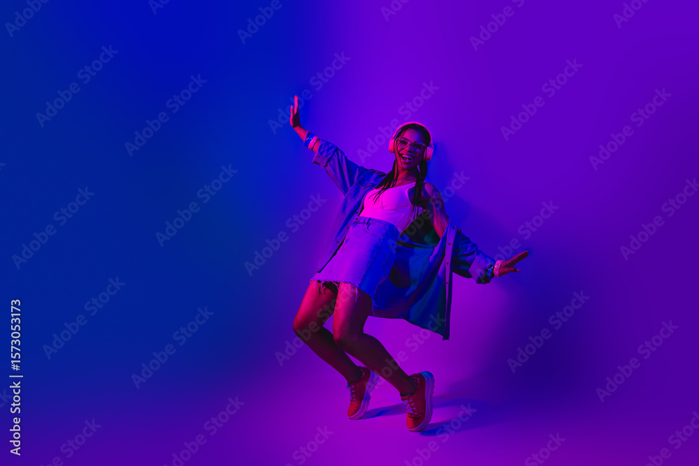 Fototapeta premium Lovely Young woman with stylish denim outfit and headphones dancing in vibrant neon light