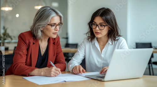 Two women analyzing documents at office, Two professional women, one older with grey hair and one younger with dark hair and glasses, are engaged in a collaborative discussion at a modern wooden table